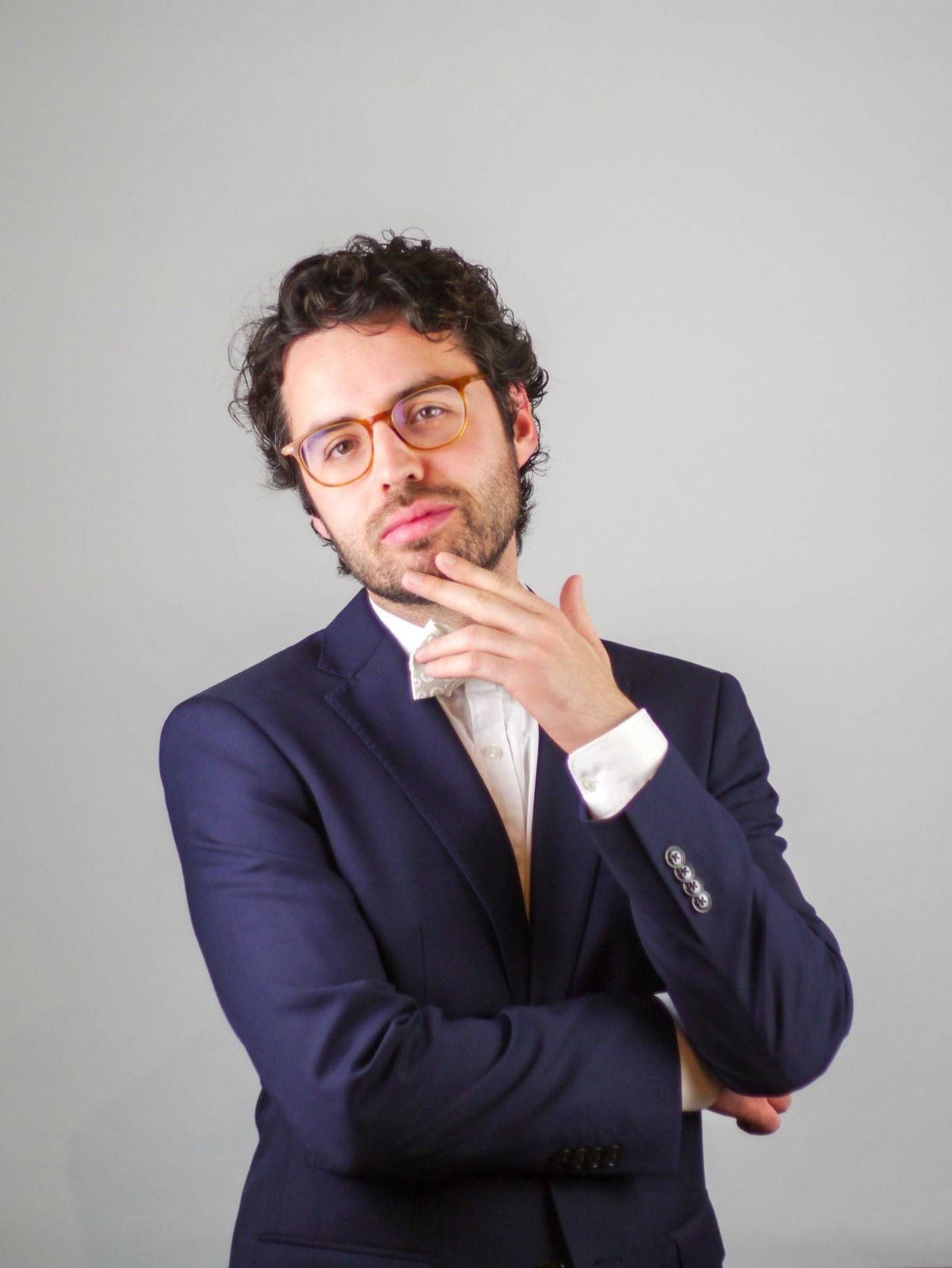 Man in glasses wearing a suit, posing thoughtfully with hand on chin against a gray background.
