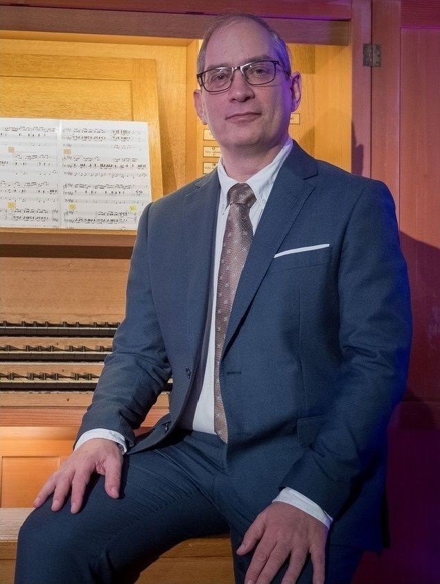 Man in a suit sitting in front of an organ with sheet music visible.