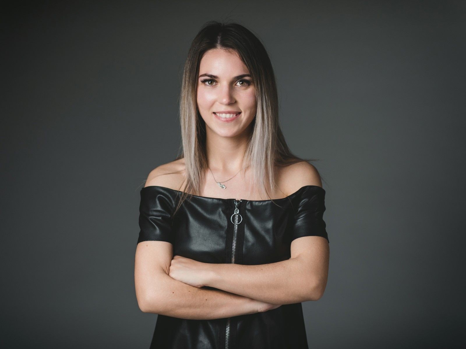 Woman in black off-shoulder top smiling with arms crossed against a dark background.